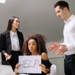 A woman in an office holds a 'HELP' sign while facing conflict with coworkers.